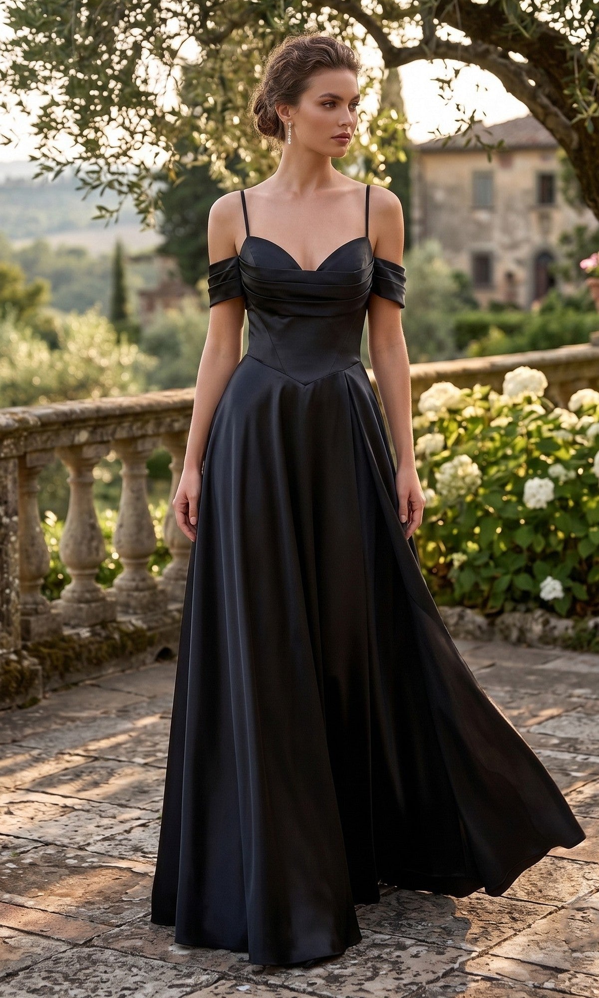 A woman wearing the Long Prom Dress: Chicas C2575 in black, featuring an elegant off-the-shoulder neckline, stands outdoors on a stone terrace surrounded by greenery and flowers.